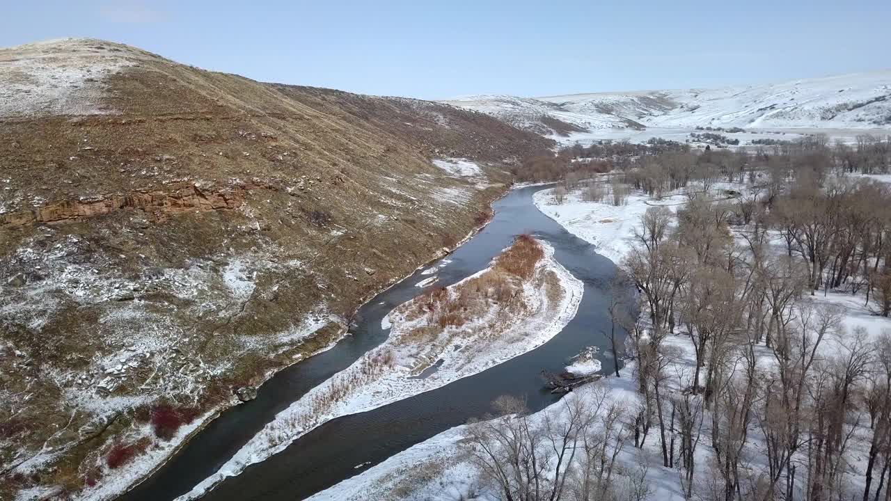 在冬季，无人机在天空的雪地上飞行，在蒙大拿比林斯，山脉和光秃秃的树木附近的河流的航拍镜头视频素材