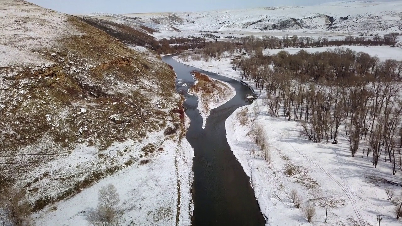 空中倾斜拍摄的河流在雪和光秃秃的树木对着天空，无人机飞越自然景观-比林斯，蒙大拿州视频素材