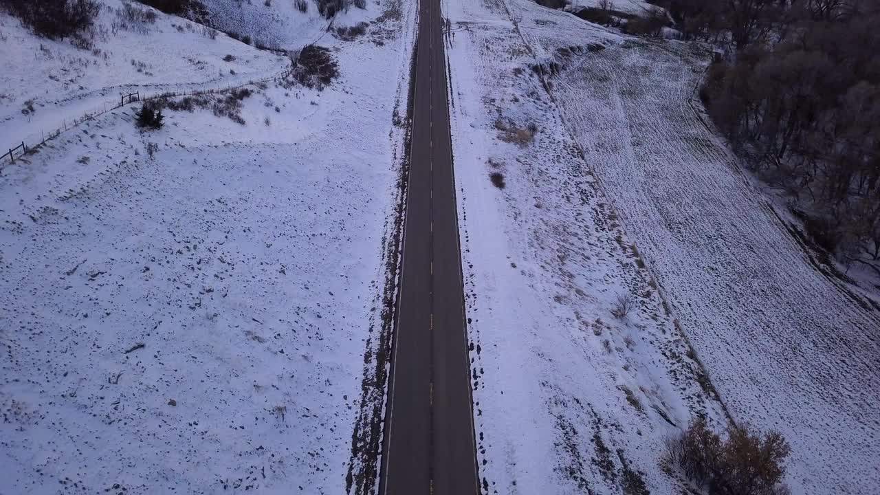 空中倾斜拍摄的空旷道路附近光秃秃的树木和山脉，无人机在日落时向后飞越雪景对天空-比林斯，蒙大拿州视频素材