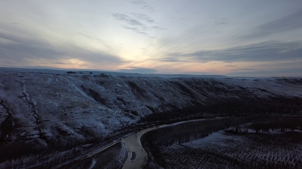 在光秃秃的树木和山脉之间的河流航拍，无人机在日落时在天空的雪景上向前降落-蒙大拿比林斯视频素材