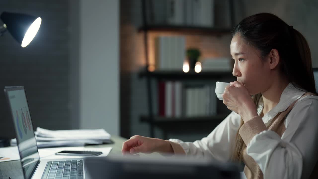 图为，一名亚洲年轻女子正在喝咖啡，以备加班时在办公桌前处理项目计划时提神。视频素材