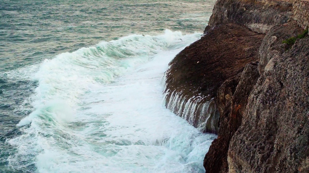 海浪冲击岩石海岸，绿色的水飞溅，形成白色的泡沫，从上面看，一些鸟飞近，慢动作视频视频素材