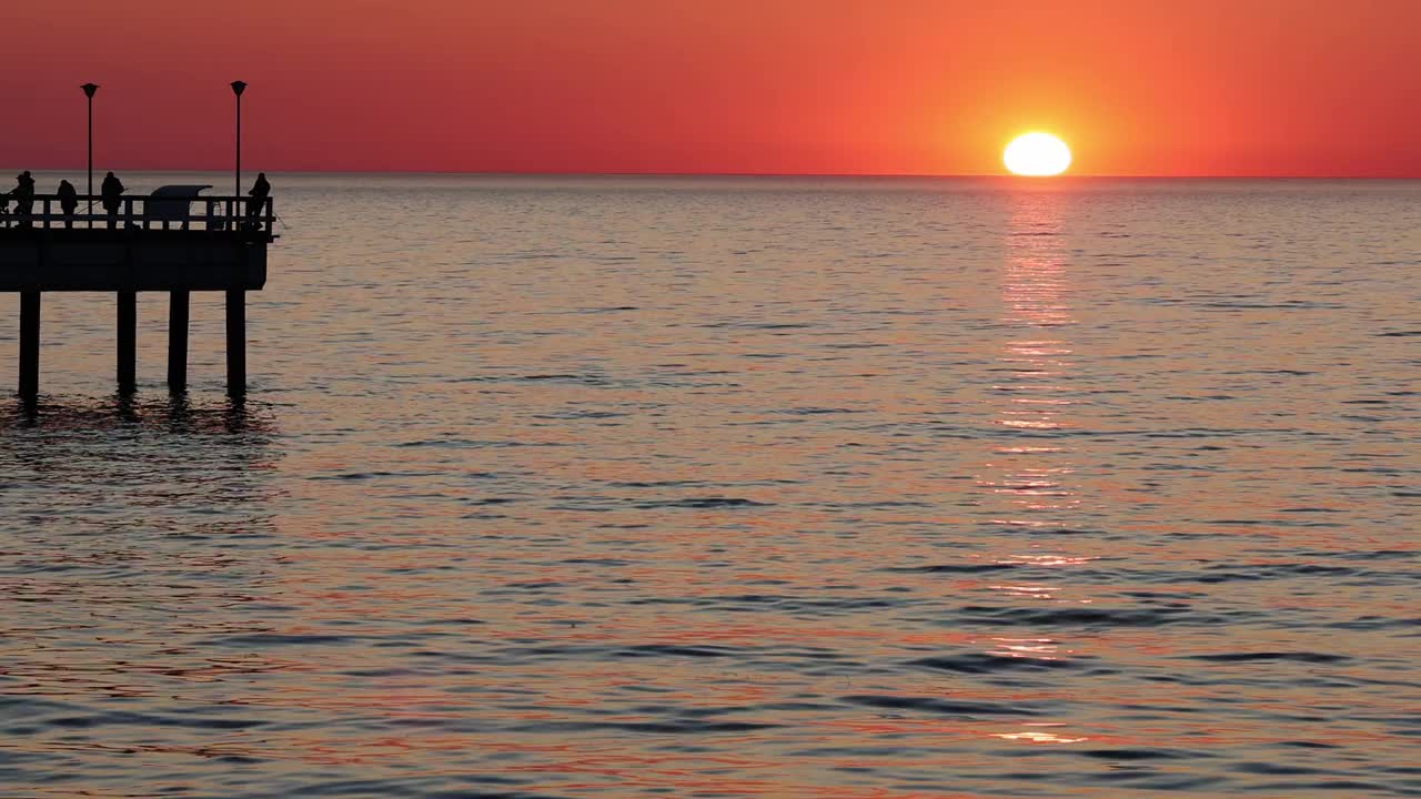 橘黄色的大太阳落在平静的海面上。渔民在码头上钓鱼。视频素材