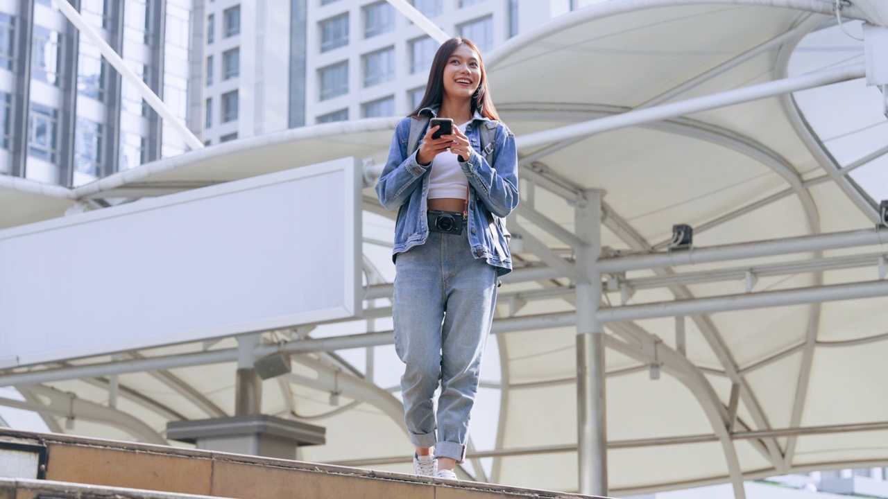 年轻的亚洲女子穿着牛仔裤，夹克，衣服，在晴朗的日子里走在城市的街道上，用智能手机发短信视频素材