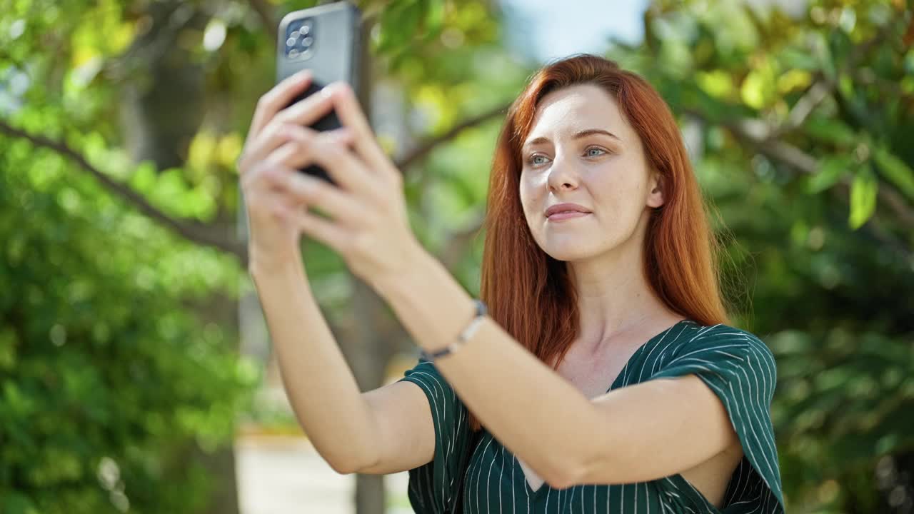 年轻的红发女子微笑着自信地在公园里用智能手机自拍视频素材