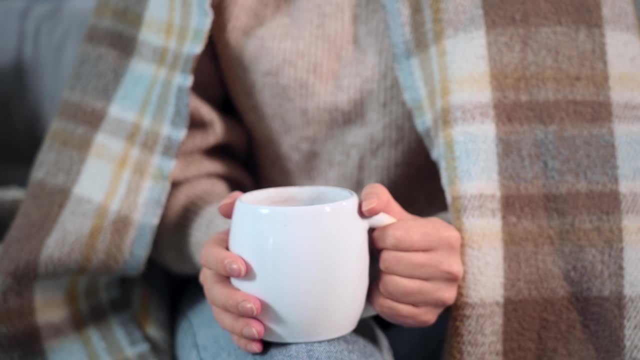 一个年轻迷人的女人坐在沙发上喝着热茶，穿着温暖的格子裙，微笑着看着镜头视频素材