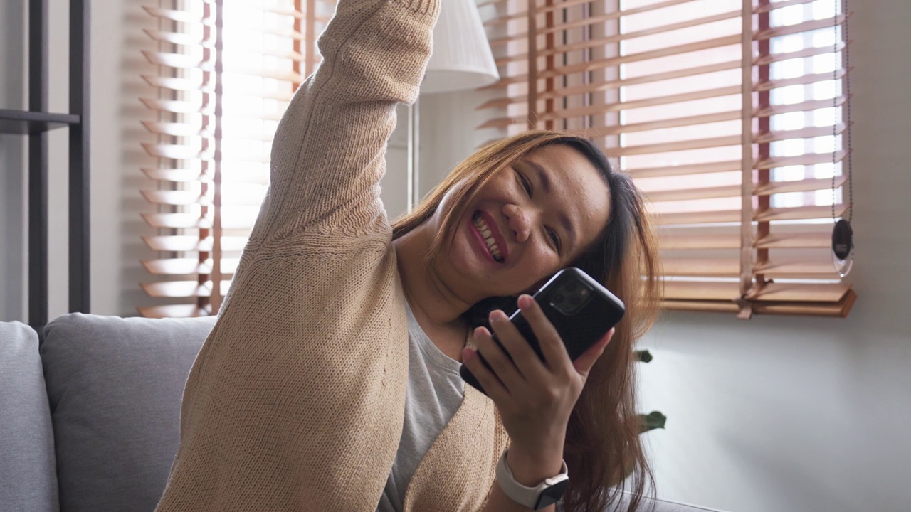 兴奋的年轻亚洲女子坐在沙发上用手机庆祝视频素材