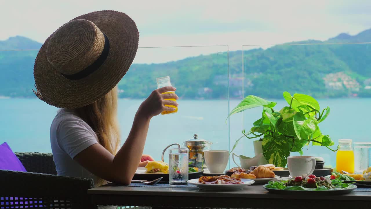 年轻女子坐在咖啡馆的桌子上，看着海景，喝着橙汁视频素材