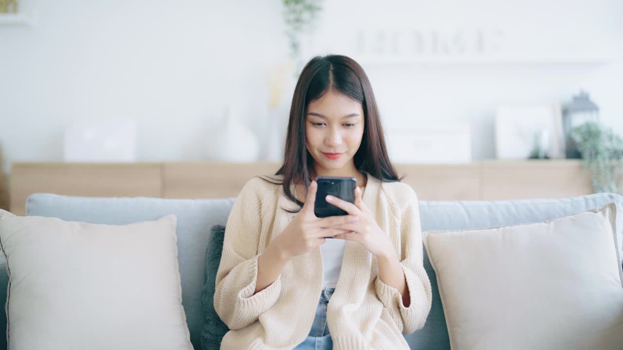 快乐的年轻亚洲女人在家里舒适的沙发上放松地用智能手机发短信，微笑的女孩用手机聊天，用小工具浏览无线互联网，在家网上购物视频素材