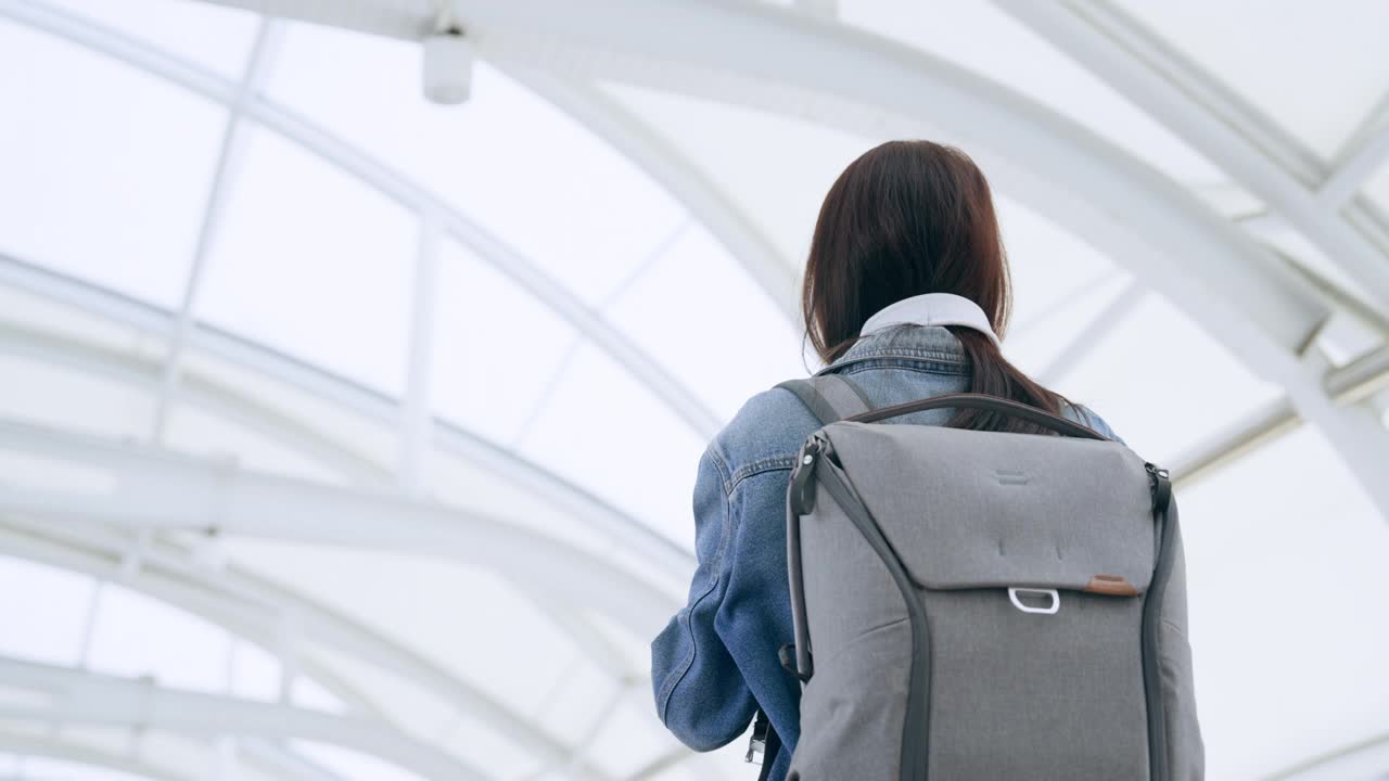 年轻的亚洲女子穿着牛仔夹克，戴着耳机听音乐，背着背包在城市里散步。青春期的女性开心，笑脸盈盈。放松和享受城市生活的年轻女士视频素材