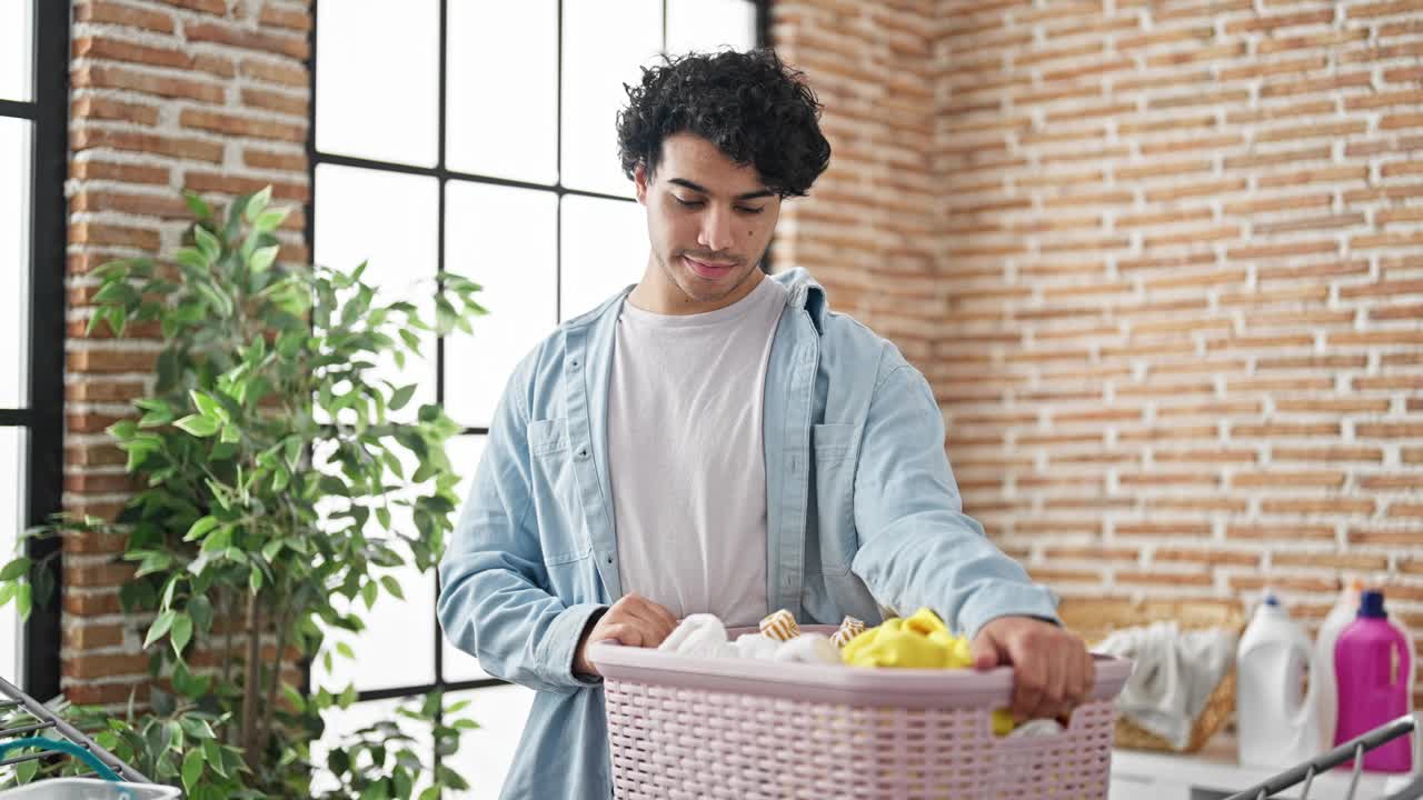 年轻的拉丁男子带着悲伤的表情在洗衣房拿着装满衣服的篮子视频素材