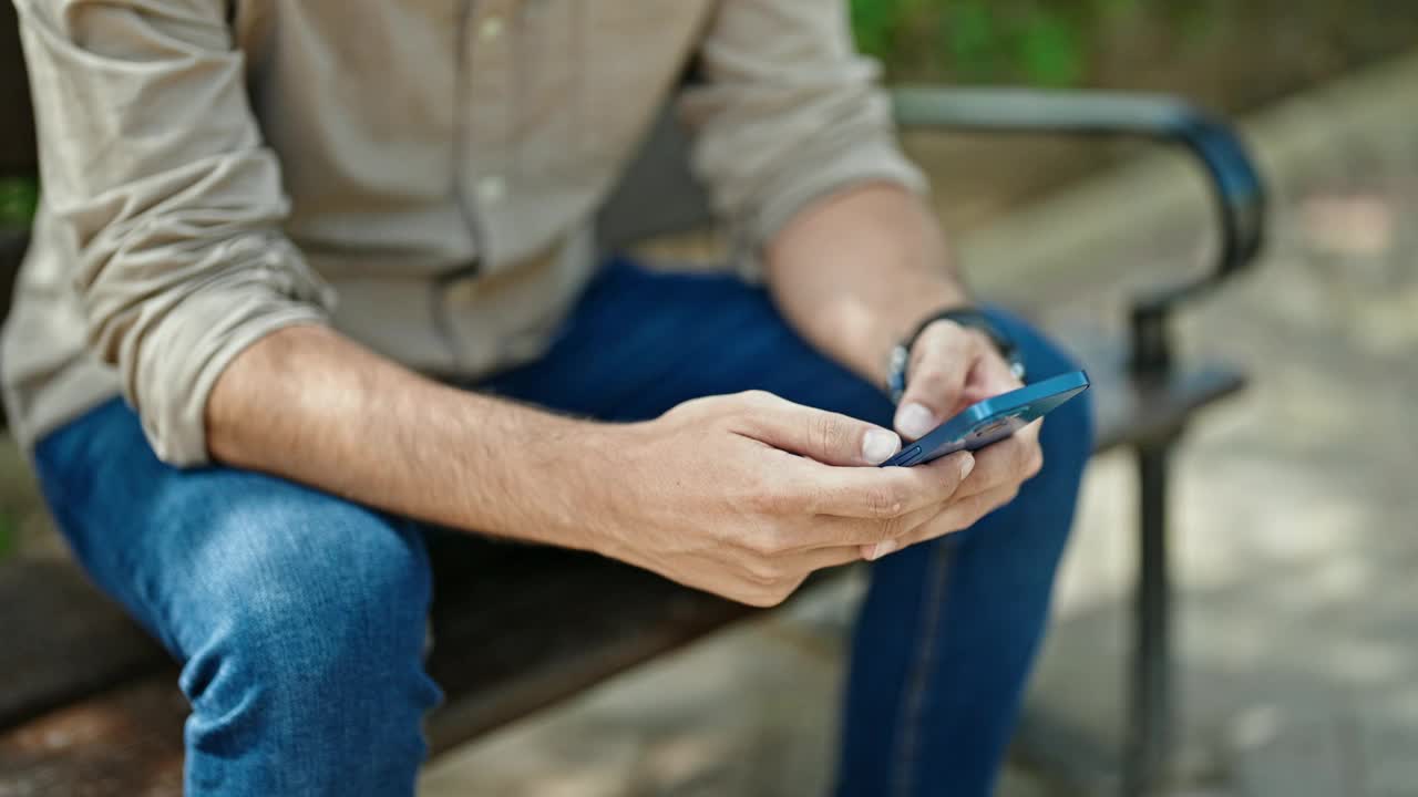 年轻的西班牙裔男子坐在公园的长椅上，表情严肃地使用智能手机视频素材