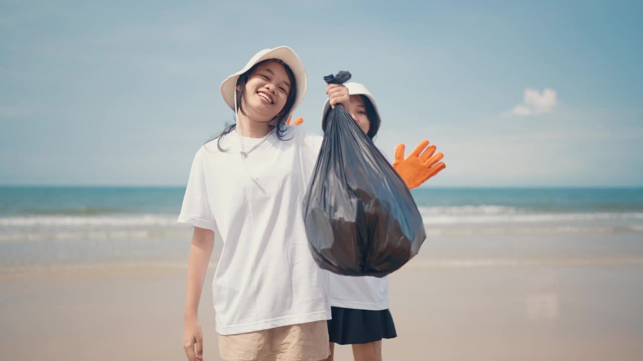 两个亚洲女孩在海滩上拿着黑色垃圾袋，开心地微笑着向镜头展示。视频下载