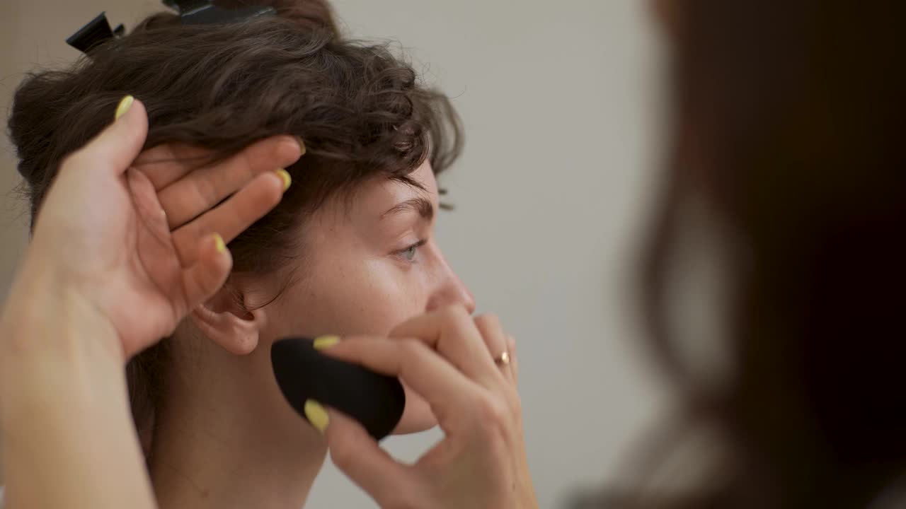 美丽的年轻女子在美容院化妆和做发型的特写。化妆师用海绵在脸上涂抹粉底液。视频素材