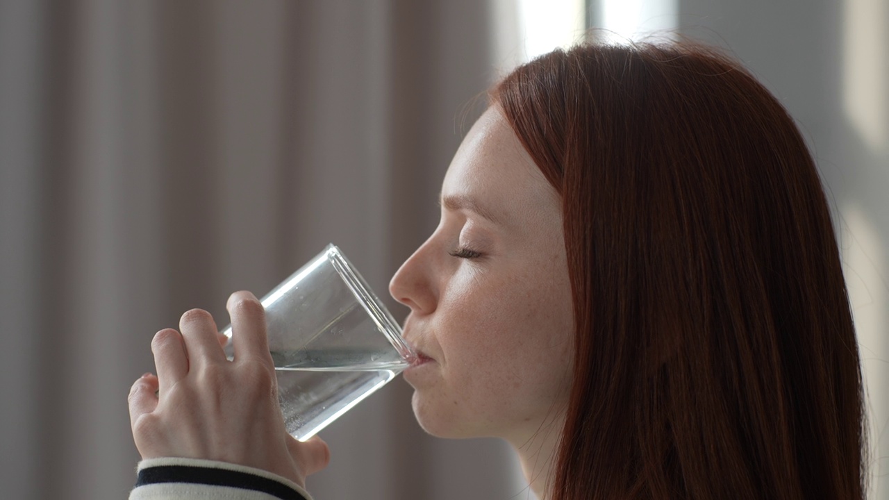 干渴的年轻红发女子的特写脸，拿着玻璃杯喝着静水，在家里享受天然的水，防止生物体脱水。视频素材