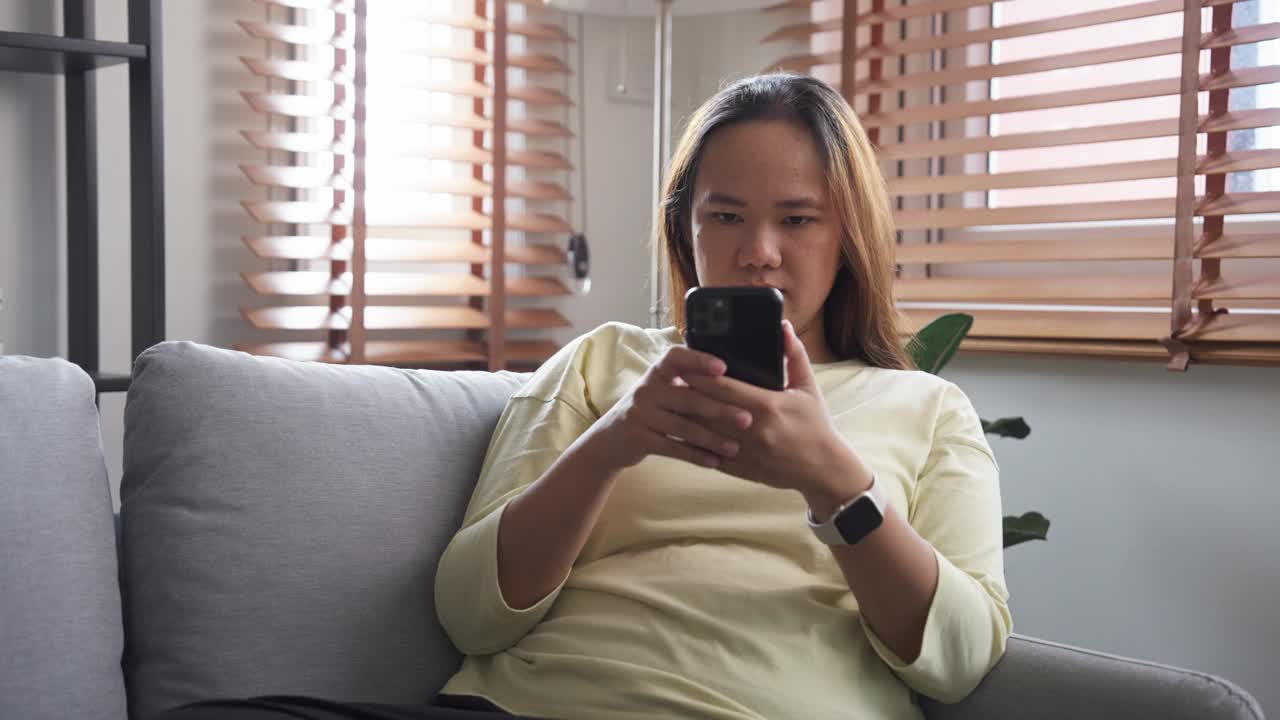 兴奋的年轻亚洲女子坐在沙发上用手机庆祝视频素材