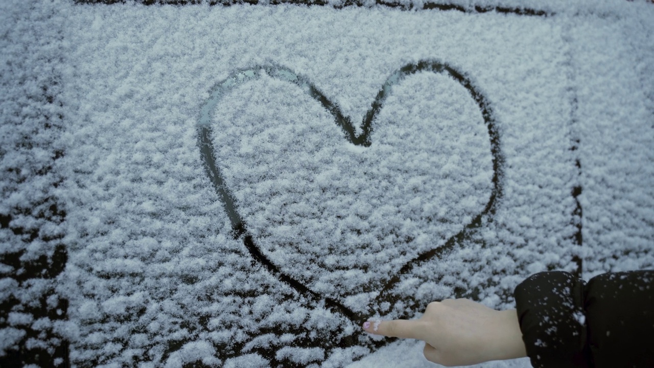 一个女孩用手指在被雪覆盖的汽车玻璃上画了一颗心。视频素材