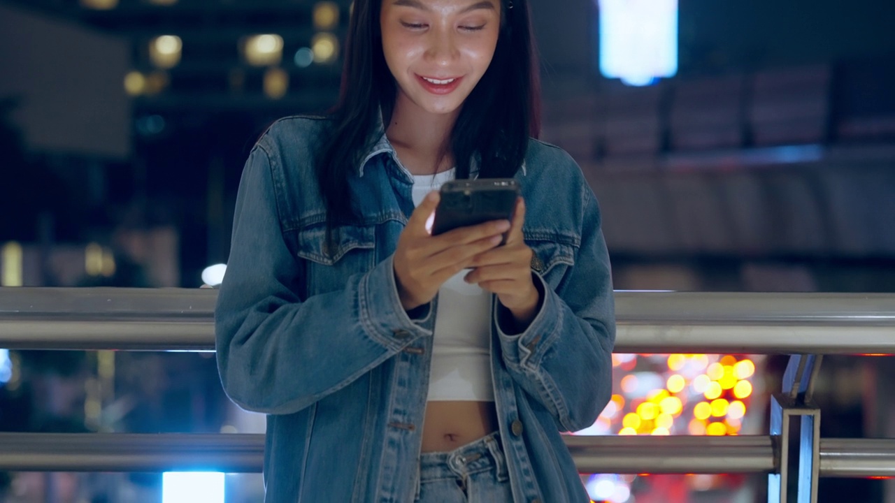 美丽的年轻亚洲女子站在城市街道在晚上使用手机。一个快乐的女人站在那里用手机打字视频素材