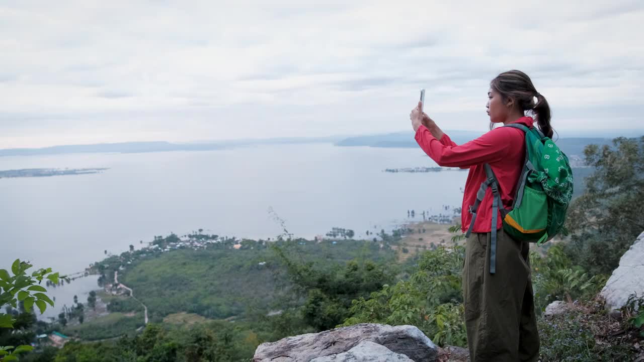 年轻的亚洲女性徒步旅行者背着背包，在山顶上用智能手机拍照，欣赏山谷湖景视频素材