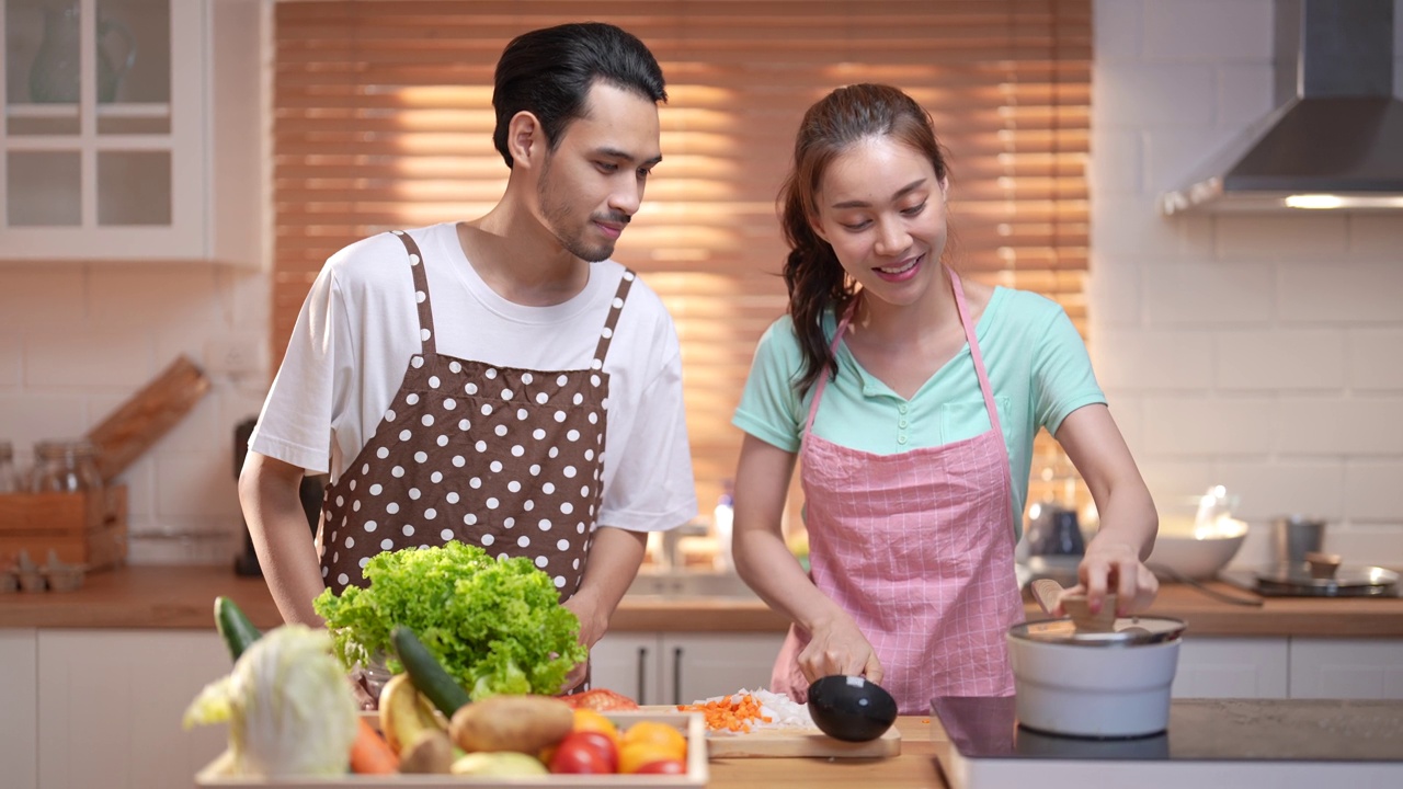 穿着围裙的亚洲年轻夫妇在厨房里一起做饭。视频素材