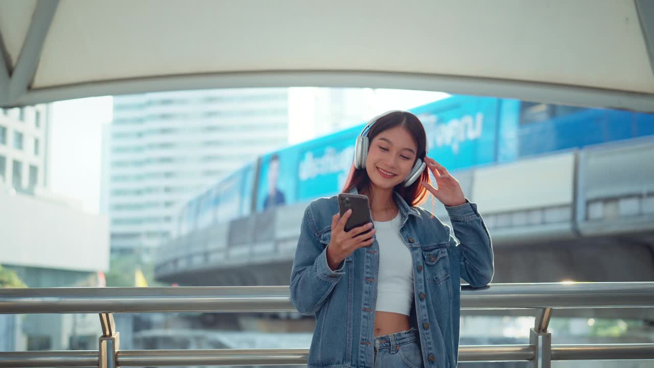 快乐的年轻亚洲女子戴着耳机在的街道上欣赏音乐，微笑的女性独自听音乐跳舞。视频素材