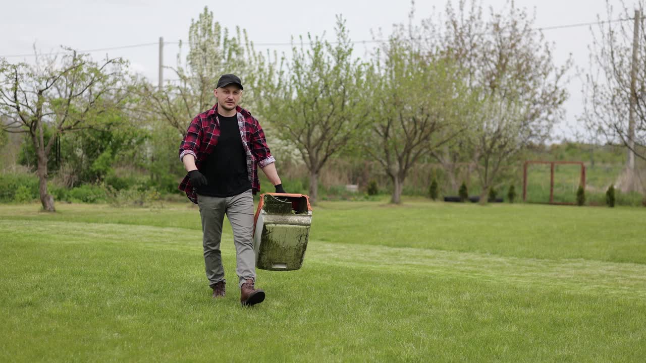春日里，穿着便装的园丁在屋后院里拿着现代电动割草机割草的塑料割草机箱。将割下的草用作堆肥或菜园视频素材