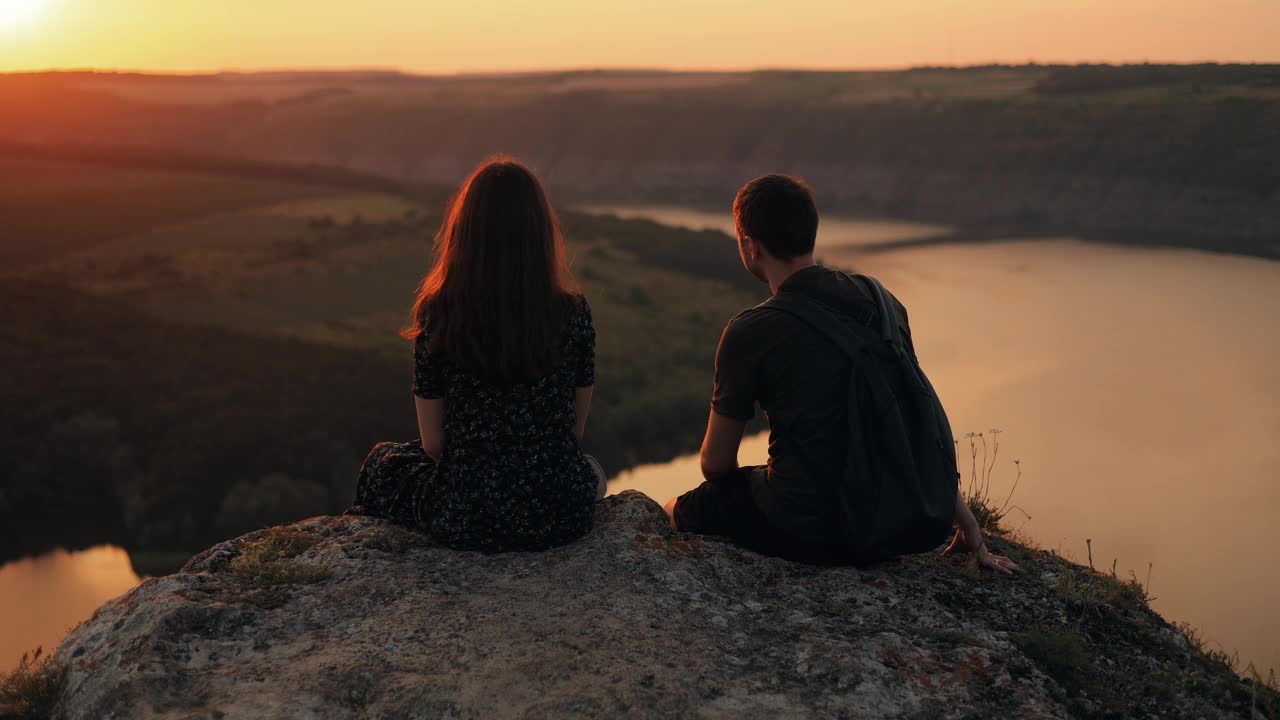 一对男女在日落时看河景，旅行中的浪漫假期。一对夫妇在河边的悬崖边看日落，在大自然中聊天，背影。视频素材