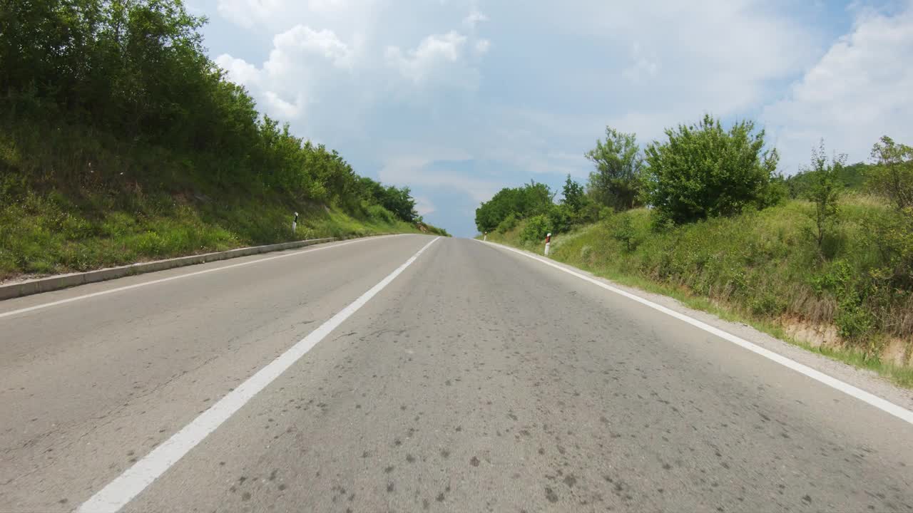 一辆汽车或摩托车沿着公路慢慢地行驶到山顶，然后下降到一个有农田的大山谷。蓝天白云。观点拍摄视频下载