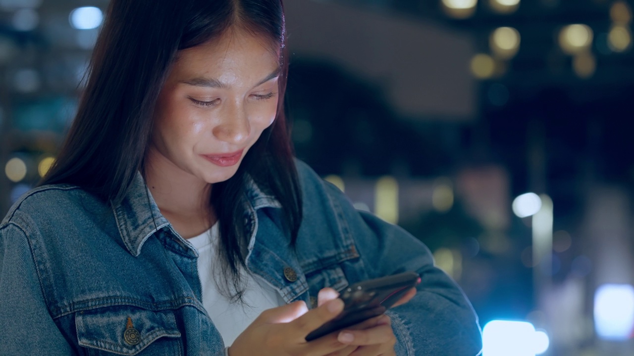 美丽的年轻亚洲女子站在城市街道在晚上使用手机。一个快乐的女人站在那里用手机打字视频素材