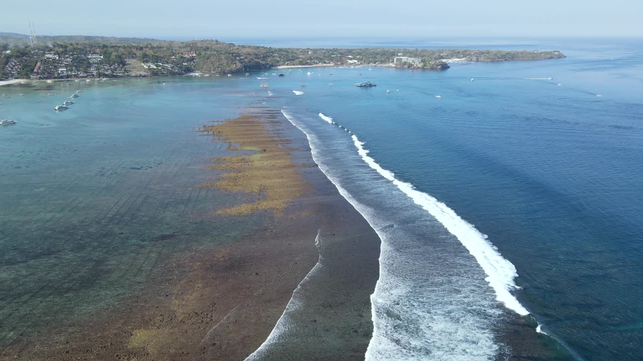 无人机拍摄的海浪到达Nusa Lembongan Jungut Batu村的海岸，海滩上有游船和海藻农场。旅游目的地靠近巴厘岛，印度尼西亚视频素材