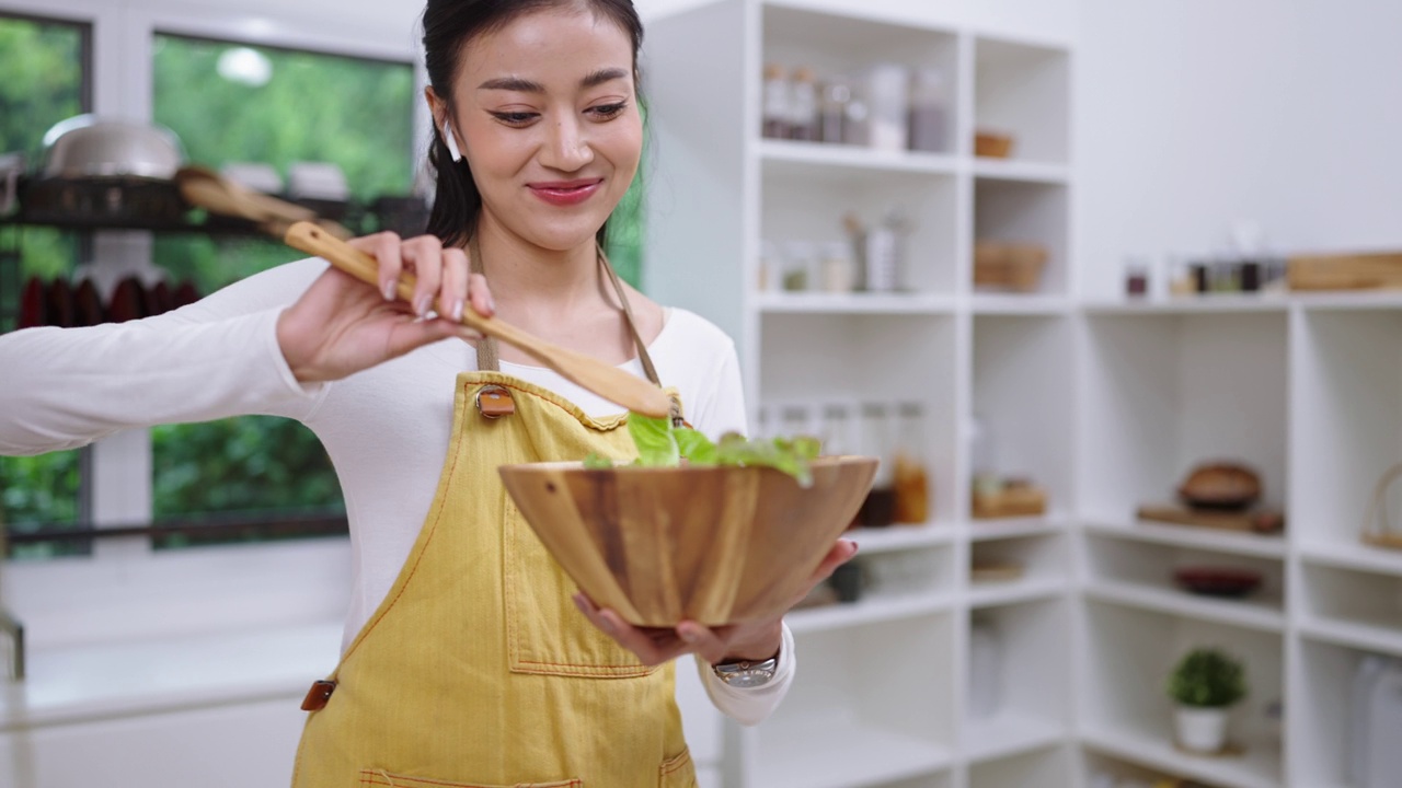 快乐的年轻亚洲女性喜欢有趣的舞蹈，拿着木制沙拉勺做饭，用叉子唱歌。女性在厨房柜台边笑边在家里准备素食健康食品视频素材