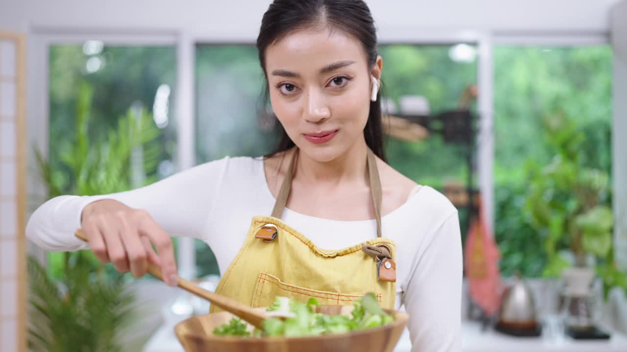 快乐的年轻亚洲女性喜欢有趣的舞蹈，拿着木制沙拉勺做饭，用叉子唱歌。女性在厨房柜台边笑边在家里准备素食健康食品视频素材