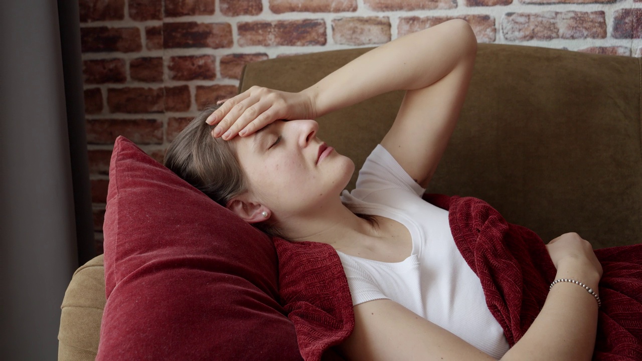 生病的头痛的女人躺在客厅的沙发上休息。保健、女性在家感到不适、疾病和健康问题。视频素材