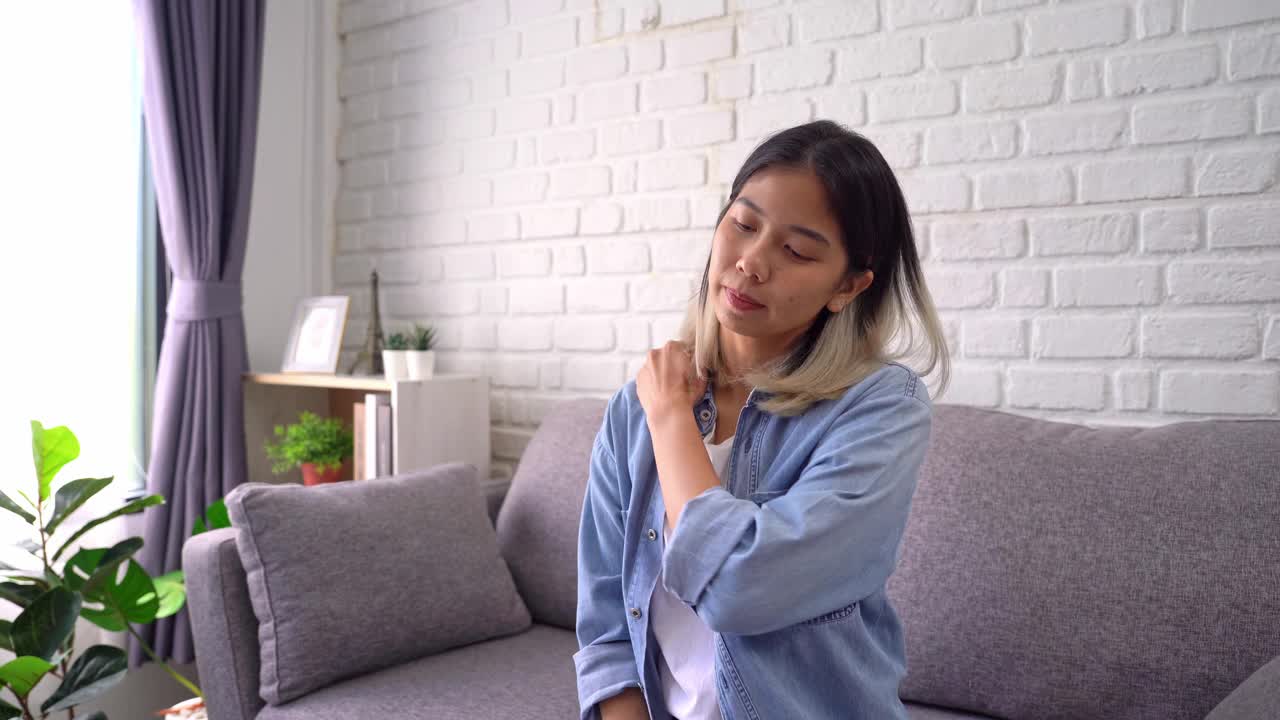 颈部疼痛的亚洲女性，她坐在家里的沙发上。视频下载