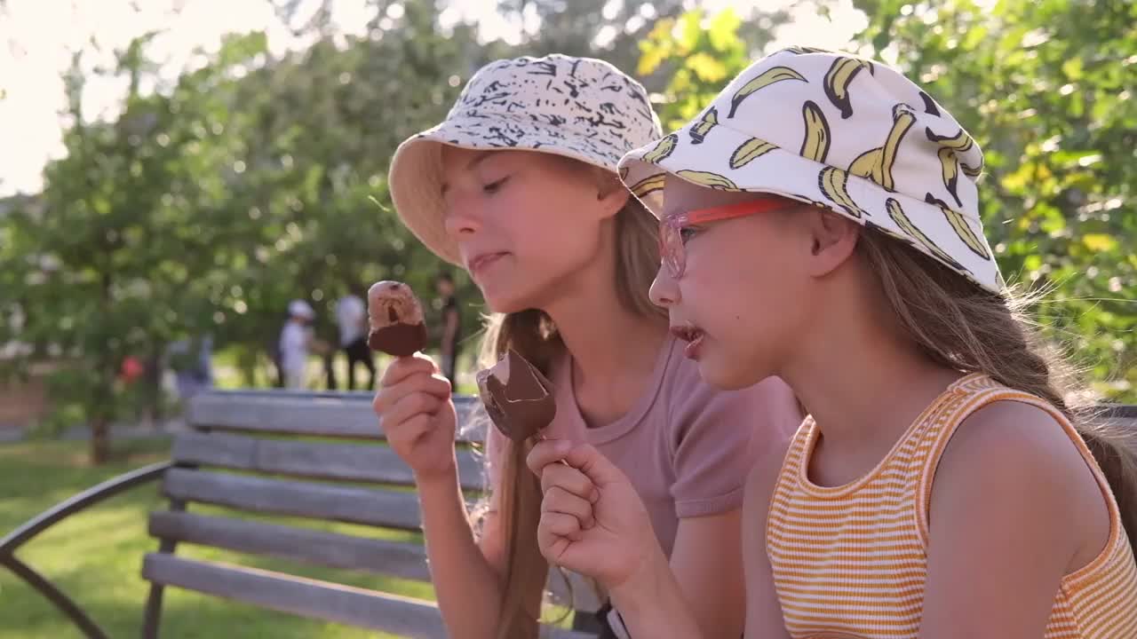 高清视频。快乐的孩子们，两个女孩，在夏天戴着巴拿马帽吃冰淇淋，坐在长椅上，在公园外面视频下载