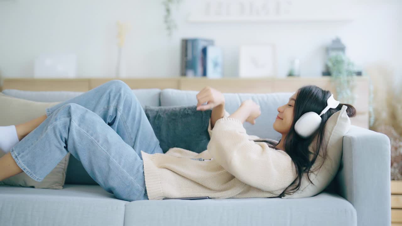 年轻的亚洲女子在家里客厅的沙发上听音乐。快乐亚洲女性使用手机，戴耳机，躺在沙发上视频素材