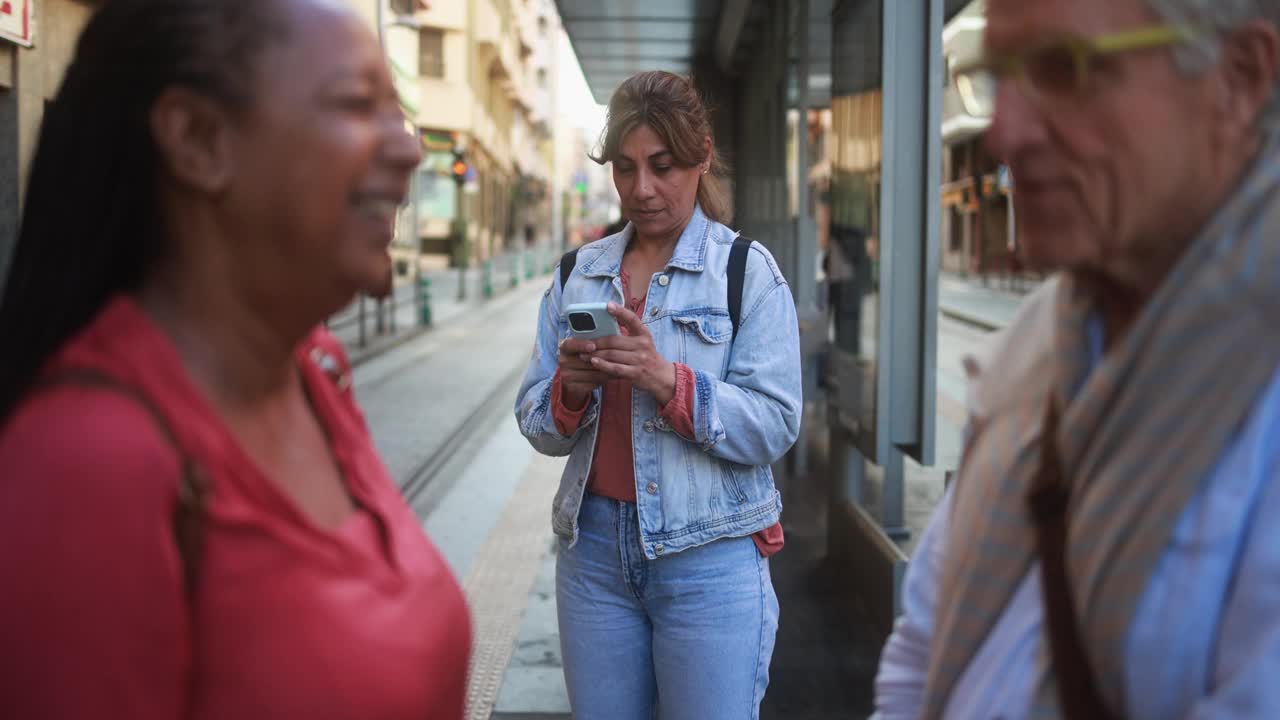 成熟的拉丁女人在电车站用手机——多种族的老人在城市里聊天视频素材