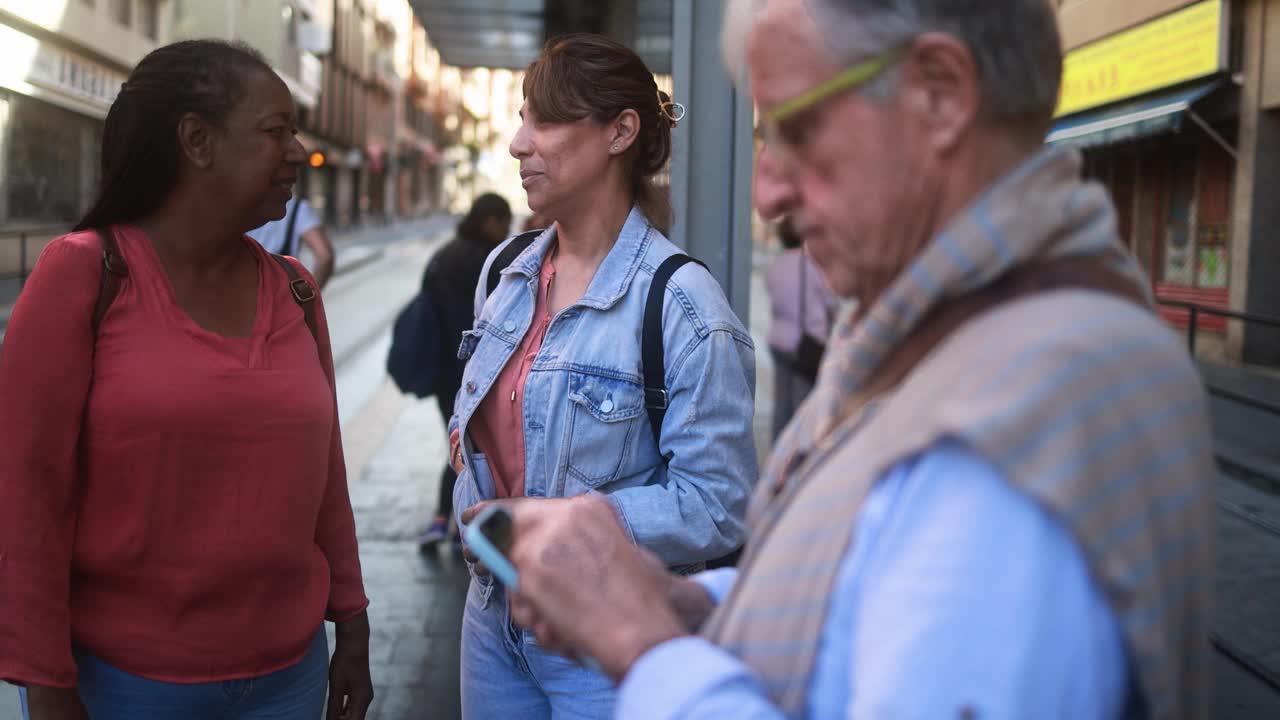 多种族妇女在电车站边等边聊天——老人在城市里用手机视频素材