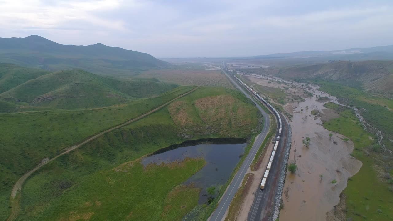 航拍的货运列车在轨道上的道路附近的河流，无人机向后飞行在景观对天空-贝克斯菲尔德，加州视频素材