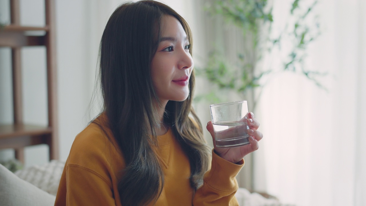 亚洲女性以健康的生活习惯开始新的一天，女性早上在家里的客厅拿着杯子喝干净的矿物质天然静水，生活方式的健康理念视频素材