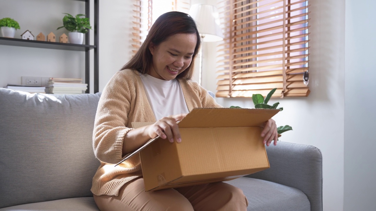 兴奋微笑的亚洲女人在家里的客厅里打开一个惊喜的盒子视频素材