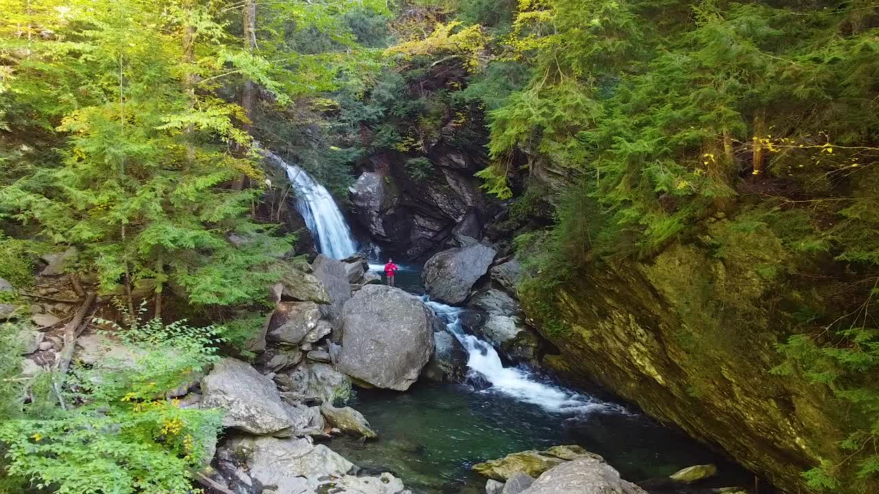 在郁郁葱葱的绿色森林的岩石峡谷中，沿着河流飞行，瀑布和孤独的徒步旅行者从巨石上观察瀑布视频素材