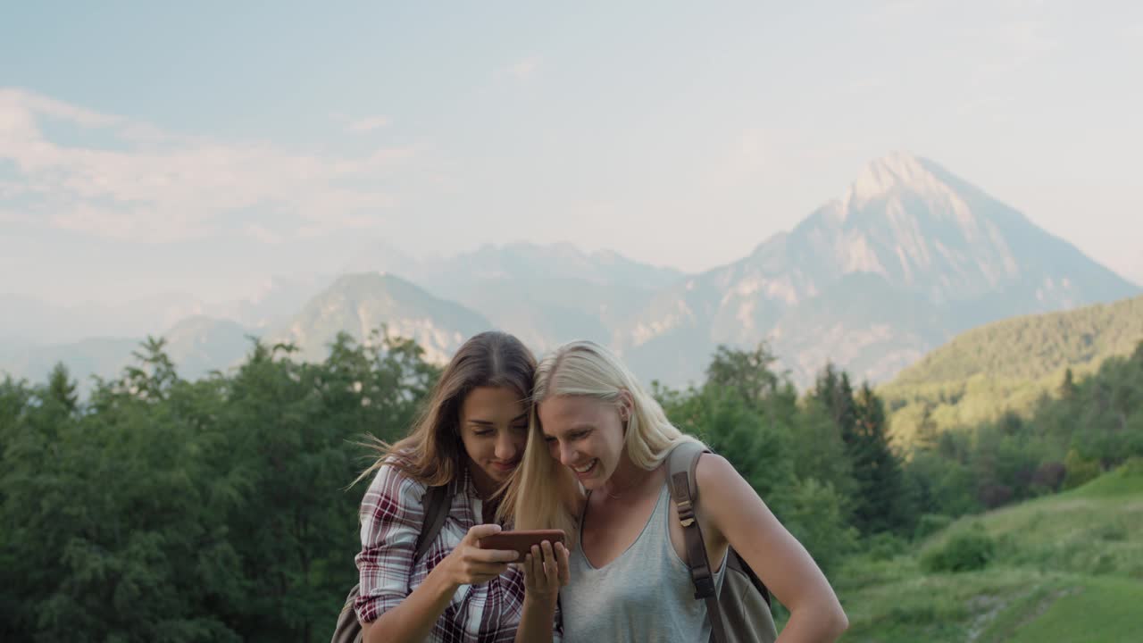 用智能手机拍照的女人最好的朋友在阿尔卑斯山徒步旅行，女孩在风景优美的自然背景下欣赏日落时的假期旅行冒险视频素材