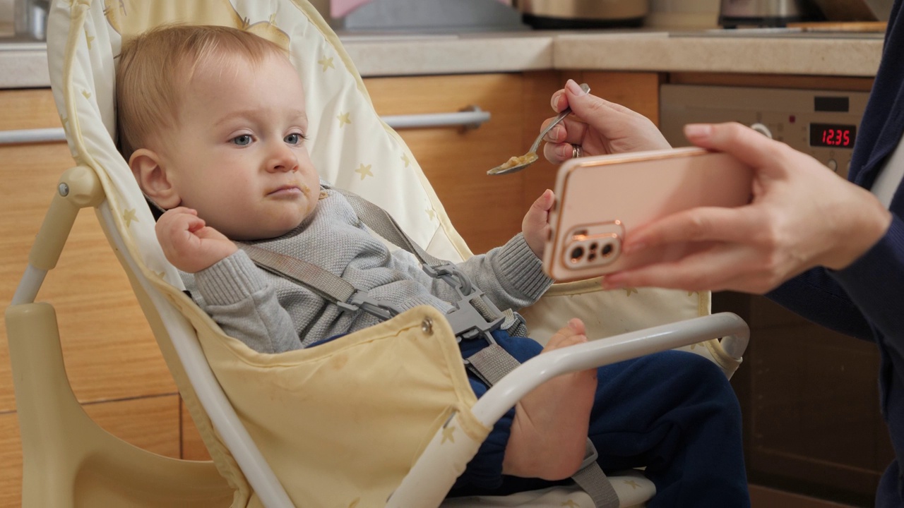小男孩坐在高脚椅上一边看动画片一边喝粥的照片。育儿理念，健康营养和婴儿喂养视频素材