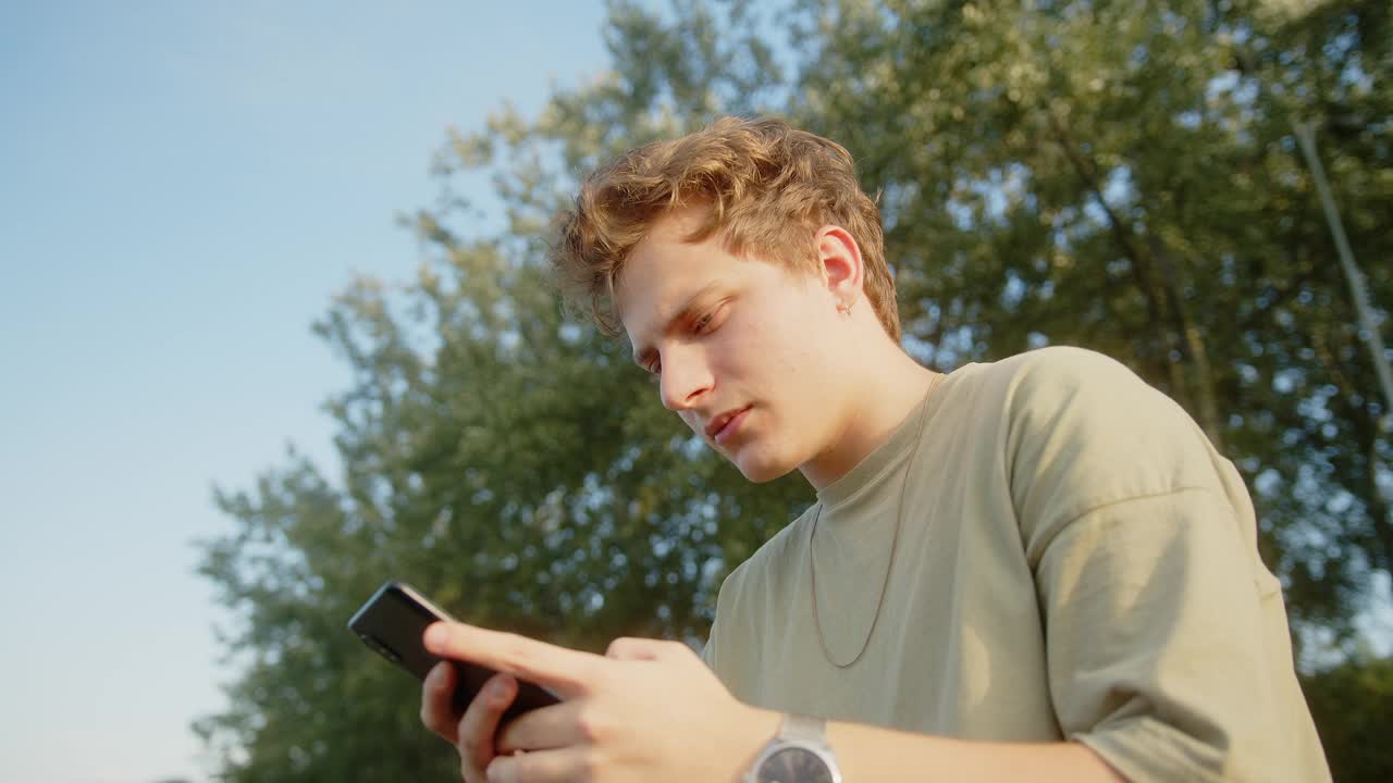 年轻的金发碧眼男孩挣扎着在智能手机上给他的朋友或女朋友发短信。美丽的自然和树木的背景视频素材