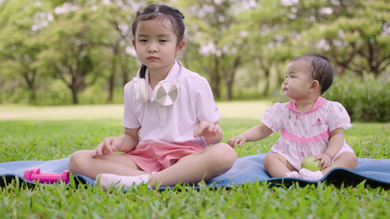 两个可爱的亚洲女孩坐在瑜伽垫上一起玩，在花园里玩得很开心。视频素材