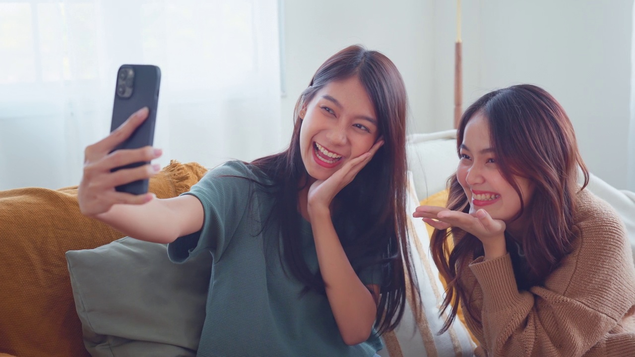 两个年轻的亚洲女人微笑着在家里的客厅用智能手机自拍。快乐的生活方式。视频素材