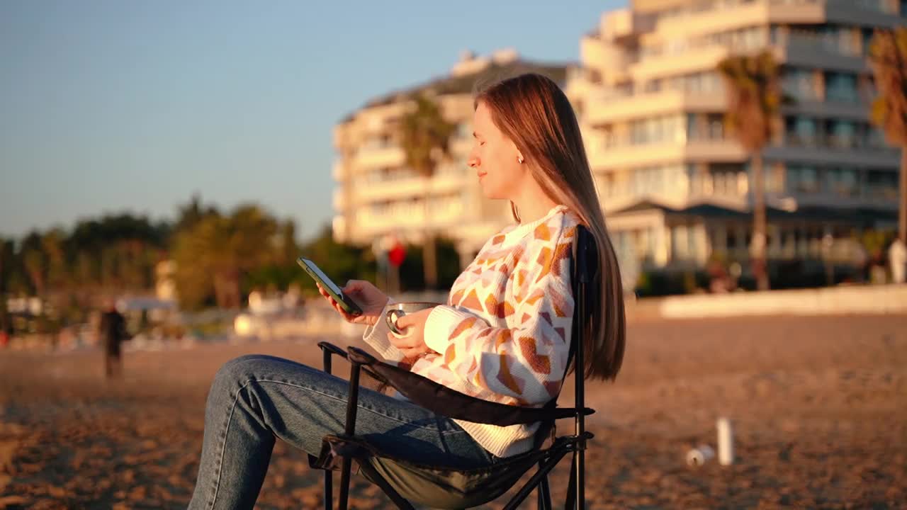 年轻漂亮的女人拿着发短信的智能手机，喝着咖啡，在冬天的海滩上，海浪的背景。可爱迷人的女孩在秋天的海边喝茶时使用手机视频下载