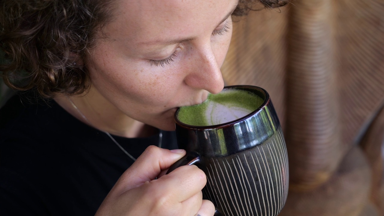 女人手里拿着一杯热腾腾的抹茶拿铁，用一杯热饮放松一整天的喧嚣。家的概念舒适的时刻与一杯热抹茶。慢动作视频视频素材