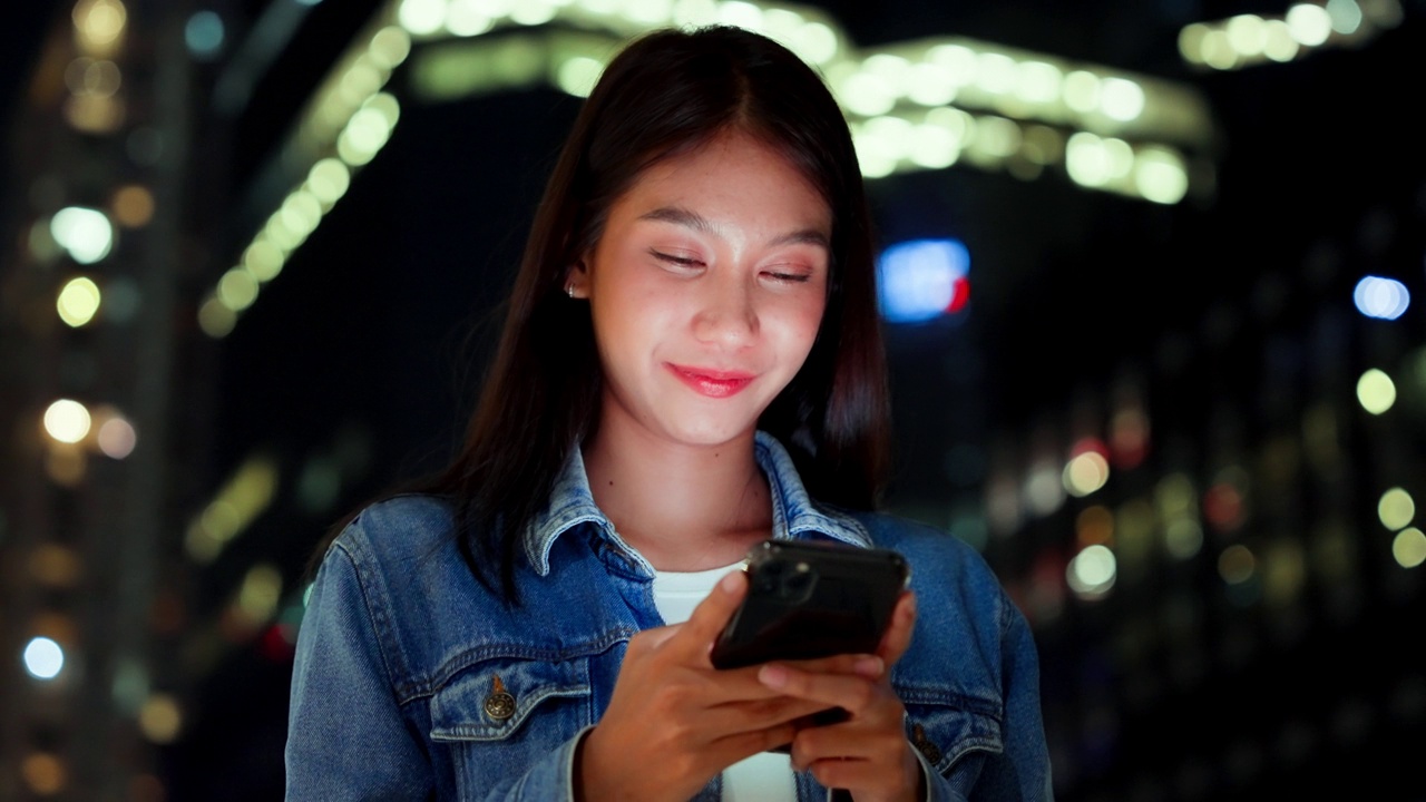 美丽的年轻亚洲女子在晚上用手机走在城市街道上。一个快乐的女人站在那里用手机打字视频素材